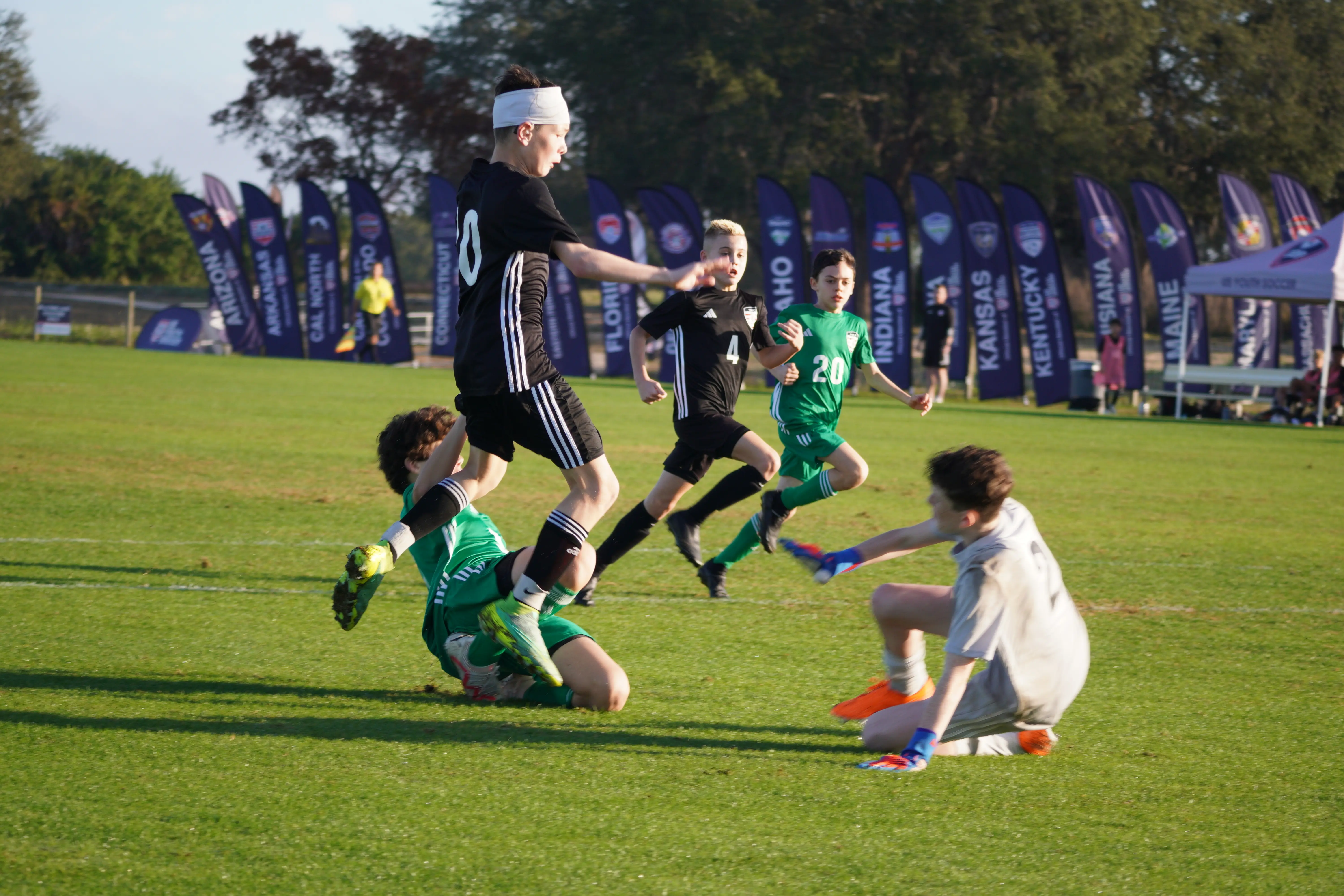 Daylen Hammon in black ODP kit with white headband jumping over sliding goalkeeper at ODP Regionals Orlando FL January 2024